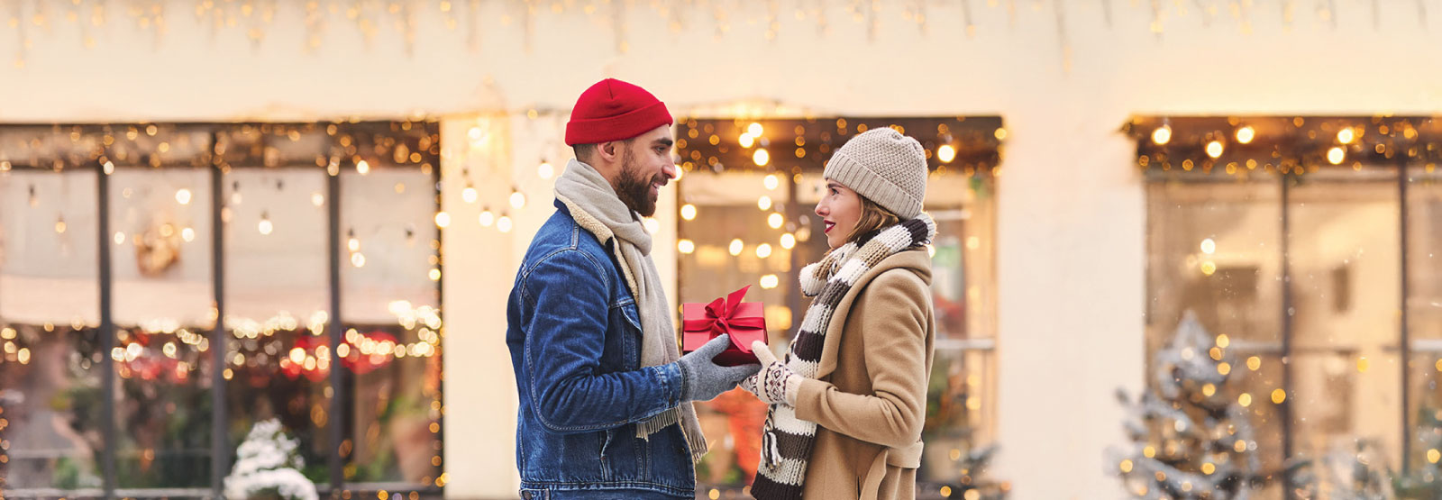 Man and woman exchanging a gift in front of stores decorated for the holidays.